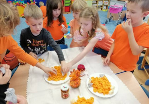 Dzieci stojąc przy stoliku degustują smakołyki w kolorze pomarańczowym.